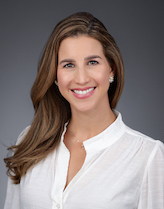 Carla Duenas smiling for a photo. She is wearing a white blouse and has a gray background with her hair down to her right side.